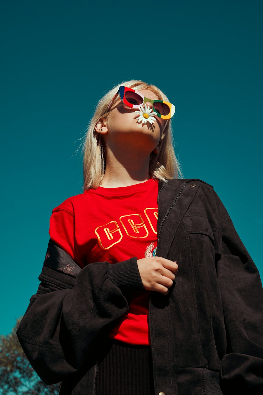 Woman in a red embroidered tee and a black suede jacket, against a teal sky