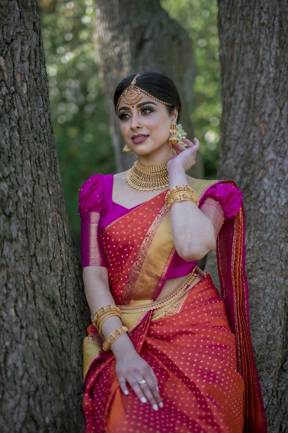 Bride in a red and pink silk saree wearing a gold choker, jhumkas and maang tikka