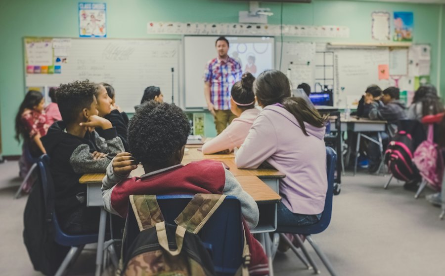 Students seated at group tables listening to their teacher at the front of the class