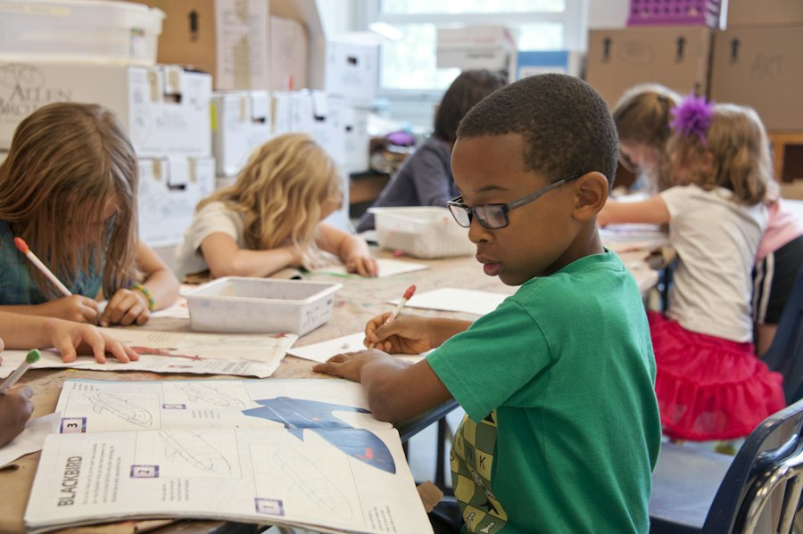 Young children writing at their desks, one boy in glasses concentrating on his work