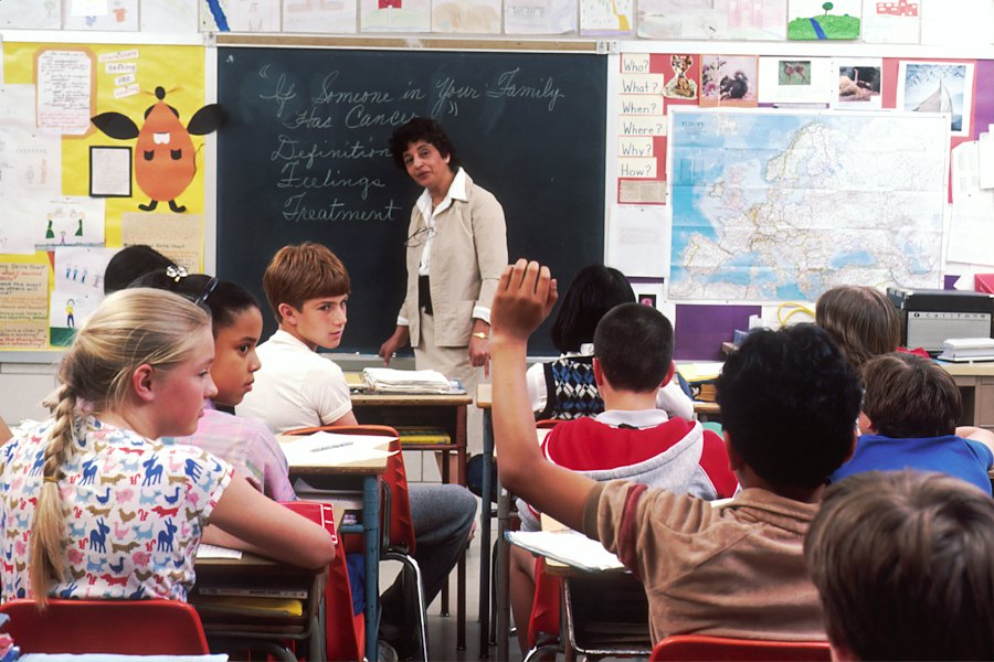 A teacher at the blackboard while students raise their hands to answer